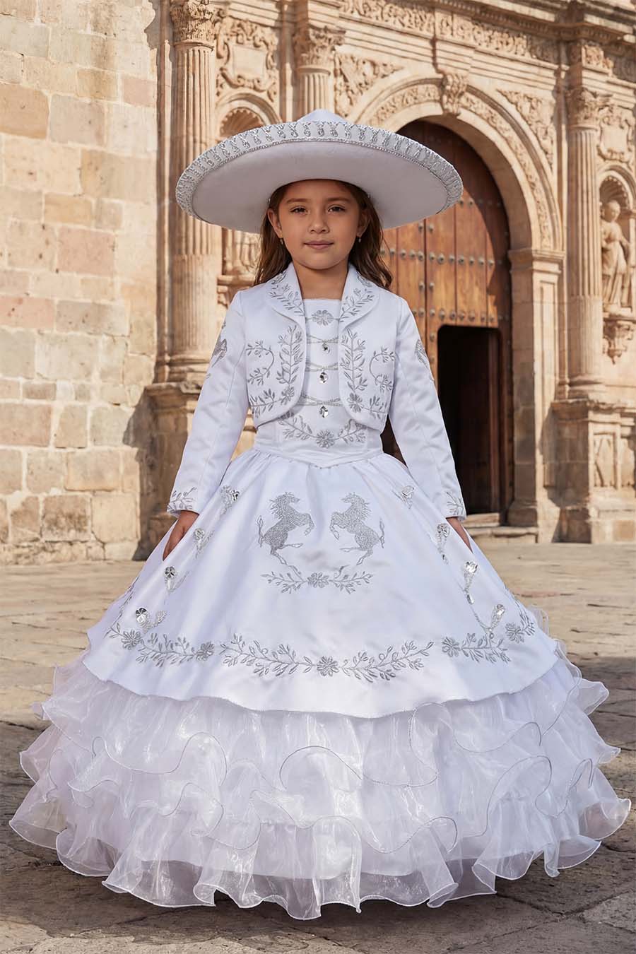 A girl wearing a traditional charro dress with silver embroidery with a sombrero in front of a colonial church