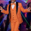 Boy wearing an rust orange tuxedo suit with black satin shawl lapels, matching vest and bow tie, dancing at a formal event with colorful party lights in the background.