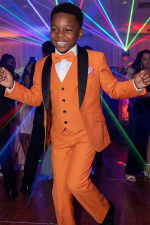 Boy wearing an rust orange tuxedo suit with black satin shawl lapels, matching vest and bow tie, dancing at a formal event with colorful party lights in the background.