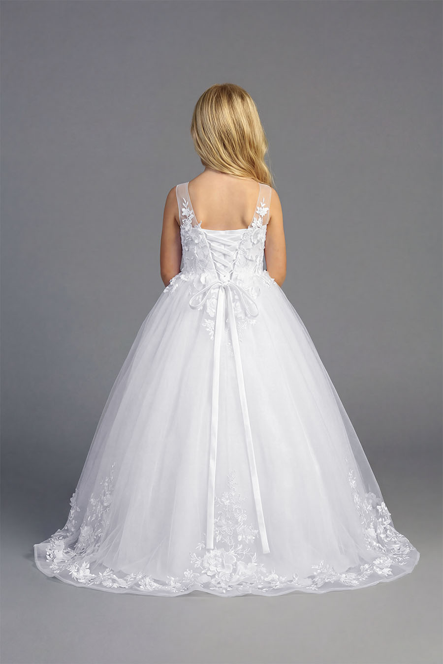 Young girl wearing a white sleeveless formal dress with floral appliqué bodice and full tulle skirt on a neutral background.